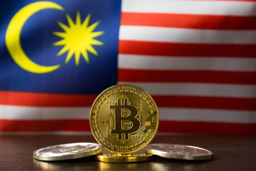 A gold Bitcoin stands upright, flanked by two silver Bitcoins, with the Malaysian flag in the background—perfect for those looking to buy crypto on Coinmama.