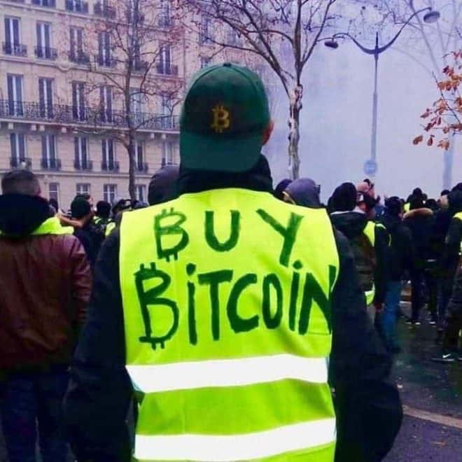 A person, in a yellow "BUY BITCOIN" vest and Bitcoin cap, promotes Coinmama amidst a crowd.